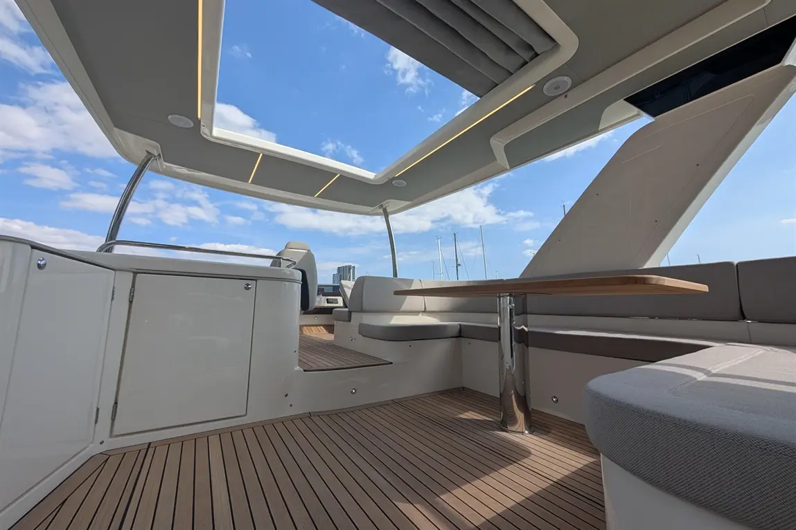 Interior view of a luxury boat with a wooden table, grey seating, and a glass ceiling, against a blue sky and marina backdrop.